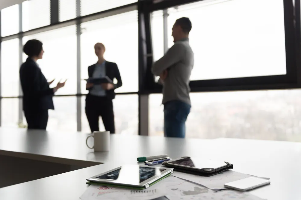 Close up of smart phone and tablet computer at office meeting room. Business people group interacting in background.